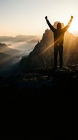Silhouette of victorious hiker with raised arms celebrating success on mountain peak at golden sunrise with dramatic landscape and misty valleys below.
