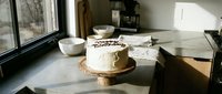 White frosted birthday cake on rustic wooden stand in sunlit modern kitchen, cozy home celebration atmosphere with natural window light.