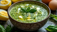 Green spinach soup with boiled eggs in rustic ceramic bowl, garnished with fresh basil leaves and cream swirls, surrounded by ingredients.