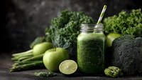 Fresh green smoothie in mason jar surrounded by organic vegetables including kale, broccoli, celery, lime and apples for healthy detox nutrition and wellness.