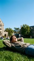 Young Caucasian male student relaxing on campus lawn with hands behind head enjoying sunny day at university college grounds with historic buildings.
