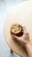 Hand holding glass of iced coffee with ice cubes on wooden round table, overhead view. Suitable for beverage, lifestyle, and cafe marketing materials.