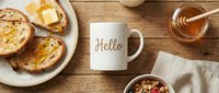White ceramic mug with Hello text on wooden table, surrounded by toast with butter, honey jar, and granola bowl for cozy morning.