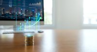 Light bulb with glowing financial growth chart and coins on wooden desk with blurred trading screens background for investment innovation concepts.