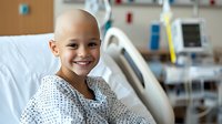 Young patient with shaved head wearing hospital gown smiles warmly while sitting in medical ward bed, showing resilience and hope during treatment.