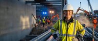 Female construction worker in safety gear smiling at industrial site with heavy machinery and workers in background during evening operations.