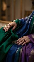Woman's hands adorned with ornate gold gemstone rings resting on vibrant silk saree in green, blue, and purple tones with golden border.