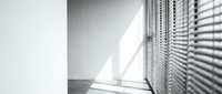 Minimalist interior corner with venetian blinds casting geometric shadows on white walls and floor creating dramatic light patterns in modern room.