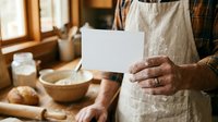 Artisan baker holding blank white card in rustic kitchen with fresh bread, flour bowl, and wooden workspace for bakery business promotion.