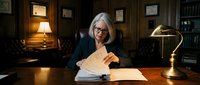Senior Caucasian businesswoman with gray hair and glasses reviewing documents at wooden desk in elegant office with warm lighting and classic decor.