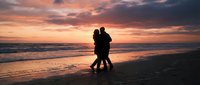 Romantic couple silhouette embracing on beach at sunset with dramatic orange and pink sky reflecting on ocean waves creating intimate moment for love concepts.