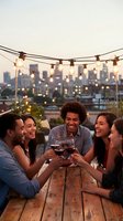 Diverse group of friends toasting with wine glasses on urban rooftop terrace at sunset with string lights and city skyline backdrop for celebration.