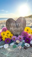 Wooden heart sign with Happy Mother's Day message surrounded by colorful flowers, seashells and sea glass on sunny beach with ocean waves and mountains.