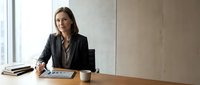 Professional businesswoman in black suit working with tablet at modern office desk near window for corporate presentations and business meetings.