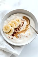 Creamy yogurt bowl with fresh banana slices, smooth peanut butter swirl and cocoa powder dusting on white ceramic plate, healthy breakfast styling.