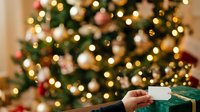 Cropped hand holding wrapped green gift box with white ribbon against blurred festive tree with golden and red ornaments, suitable for holiday marketing and seasonal promotions.