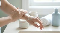 Woman applying white moisturizing cream on hands in bright bathroom with skincare products for daily beauty routine and self-care wellness.