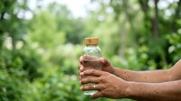 Person holding a water bottle outdoors in nature with green foliage background, suitable for hydration, wellness and healthy lifestyle concepts.