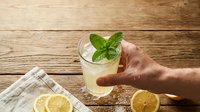 Fresh lemonade with mint in glass held by hand on rustic wooden table. Summer refreshing drink with lemon slices and striped napkin for beverage concepts.