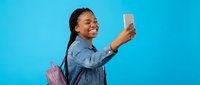 Happy young African American woman taking selfie with smartphone wearing denim jacket and pink backpack on bright blue background for social media content.