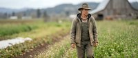Mature male farmer in casual work attire standing in vegetable field with rural farmland and mountains in background, suitable for agriculture and farming industry content.