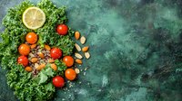Fresh organic kale leaves arranged with cherry tomatoes, lemon slice, and scattered almonds on dark green textured background, food photography layout.