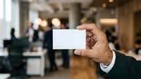 Businessman holding blank white card in modern office lobby with blurred colleagues in background, suitable for corporate branding and business communication concepts.
