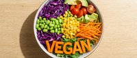 Colorful vegan bowl with fresh vegetables, corn, edamame, carrots, red cabbage, and tomatoes, topped with letter tiles spelling VEGAN on wooden table.
