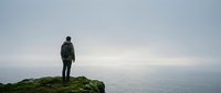 Person standing on mossy cliff edge overlooking misty ocean horizon, contemplative solitude and adventure travel concept for wanderlust and exploration themes.
