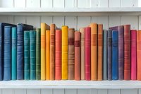 Colorful vintage books arranged in rainbow order on white wooden shelf, spines showing vibrant spectrum from blue through orange to red. Library collection display.