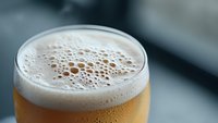 Close up view of fresh draft beer in glass with creamy foam head and condensation droplets on surface, golden amber color against blurred background.