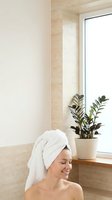 Young Caucasian woman with white towel on head smiling in modern bathroom with natural light and green plant on windowsill for spa wellness routine.
