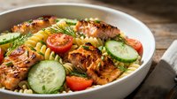 Grilled salmon with pasta salad featuring cherry tomatoes, cucumber slices and fresh dill in white bowl. Healthy Mediterranean meal for lunch or dinner.