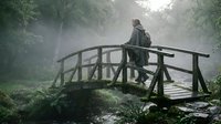 Middle-aged man with light skin tone wearing outdoor gear standing on wooden bridge in misty forest stream environment, suitable for adventure, nature, travel and lifestyle editorial use.