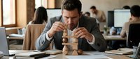 Focused businessman carefully stacking wooden blocks at office desk, demonstrating strategic planning and problem-solving skills in corporate environment.