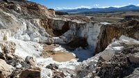 Dramatic limestone quarry landscape with white rock formations and muddy pool, surrounded by rugged cliffs against mountain range backdrop and blue sky.