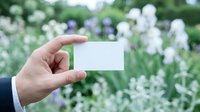 Hand holding blank white business card mockup against blurred green garden background with purple flowers for branding and corporate identity design.