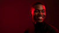 Young African American man smiling confidently in dramatic red lighting against dark background for portrait photography and creative projects.
