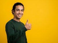 Young man with dark hair and olive complexion wearing dark green long-sleeve shirt giving thumbs up gesture against bright yellow background, expressing approval and positivity.