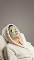 Woman in white bathrobe relaxing with green clay facial mask and cucumber slices on eyes during spa treatment for skincare and wellness routine.