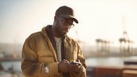 African American man in brown jacket and cap checking smartphone at harbor during golden hour sunset with industrial port background.
