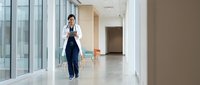 Asian female doctor walking confidently through modern hospital corridor wearing white coat and stethoscope in bright medical facility.