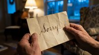 Elderly hands holding a vintage paper note with the word Sorry written in elegant calligraphy, warm home interior background.
