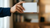 Male hand in dark blue sleeve holding blank white business card against blurred office background, suitable for corporate branding and business identity mockups.