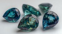 Luxurious teal and blue gemstones with pear-shaped facets arranged on neutral background, showcasing brilliant cuts and natural color variations for jewelry design.