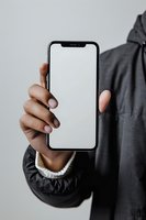 Person with dark skin tone holding smartphone with blank white screen against neutral background, wearing gray jacket.