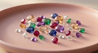 Collection of colorful precious gemstones including emeralds, sapphires, rubies and diamonds scattered on elegant pink ceramic plate for jewelry making.