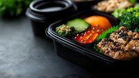 Healthy meal prep container with grilled chicken, red caviar, fresh vegetables and broccoli. Black plastic bento box on dark background for food delivery.