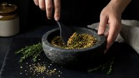Hands grinding coriander seeds in stone mortar and pestle with fresh herbs on dark background for cooking and spice preparation.