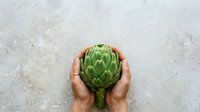 Fresh artichoke held in hands on stone surface, suitable for food, cooking, agriculture, and healthy eating content.